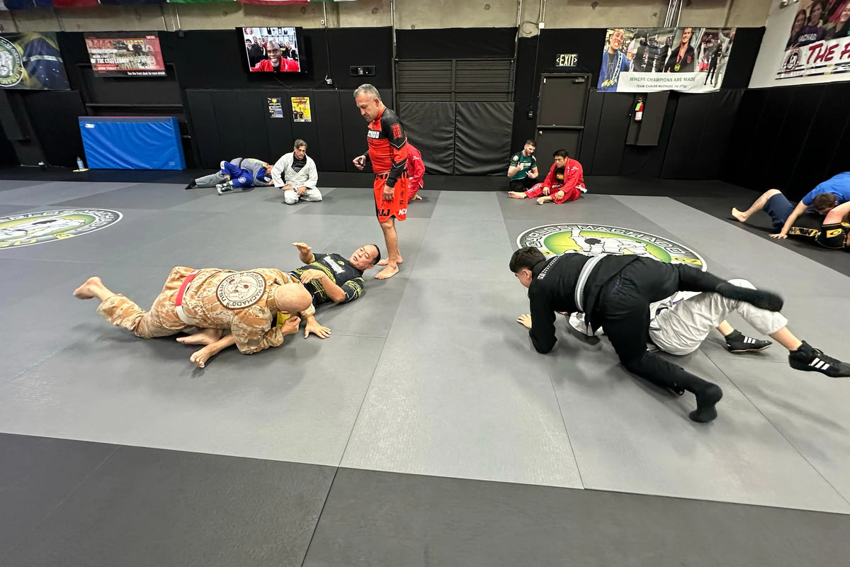 Two pairs of adults rolling on the mat at the academy, with a coach walking between them.